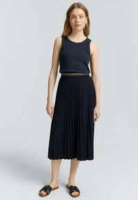 Gonna midi plissettata blu navy abbinata a un top senza maniche dello stesso colore. Presenta un leggero accento dorato in vita. Indossata con sandali neri a fascia.