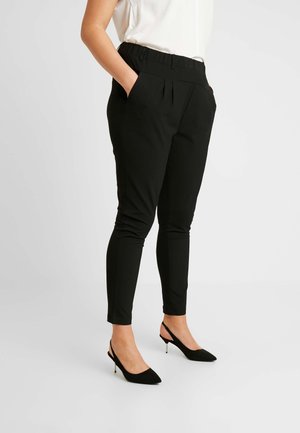 Kvinde iført tætsiddende sorte ankellange bukser og sorte peep-toe slingback hæle med en hvid top, stående med hænderne i lommerne.