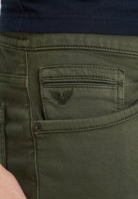 Olijfgroene denim broek heeft een klein ritszakje en een geborduurd vleugellogo op de achterkant. Glad materiaal met contrasterende stiksels.