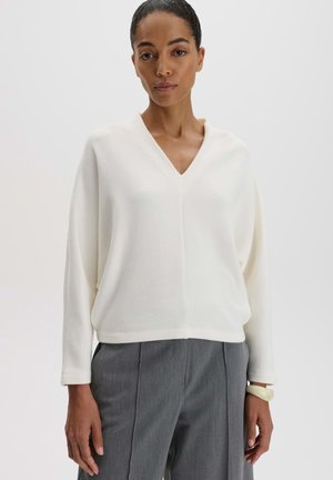Woman wearing a white V-neck long-sleeve top and gray trousers, standing with neutral expression against a plain light background.
