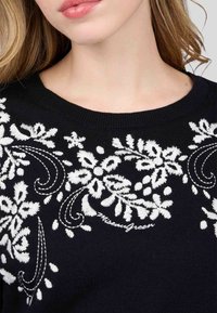 Pull noir avec broderie florale blanche et motifs tourbillonnants, accompagné de texte. Encolure ronde, texture tricotée et coupe ajustée.