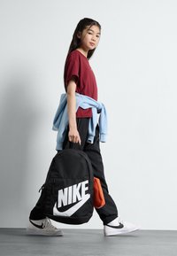 Juodas Nike kuprinės su balta logotipu, oranžiniu akcento kišeniu, derinamas su raudonu marškinėliu, mėlynu megztiniu aplink liemenį, juodomis kelnėmis ir baltomis sportbatėmis.