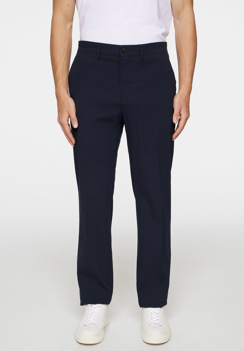 Trousers navy blue yang dirancang dengan bahan ringan, menampilkan potongan ramping, saku samping, dan detail lipatan depan, dipadukan dengan sepatu sneakers putih.