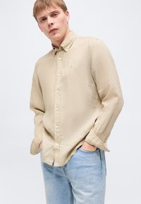 Νέος άνδρας φορώντας ένα μπεζ πουκάμισο με κουμπιά και ανοιχτό μπλε τζιν, στέκεται με τα χέρια στις τσέπες σε απλό φόντο.