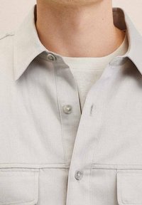 Camisa gris claro con botones, cuello abierto, dos bolsillos en el pecho y textura suave. Presenta cierres de botones y detalles de costura sutiles.