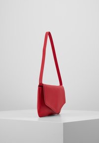 Červená kožená crossbody taška s klopou, zavěšená na šedém pozadí, vystavená na bílé ploše.