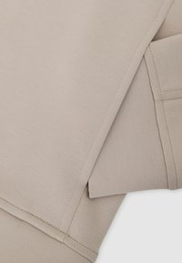 Beiger Sweatshirtstoff, glatte Textur, mit umgeschlagenem Bündchen und sichtbaren Nähten. Einfaches Design ohne zusätzliche Muster oder Akzente.