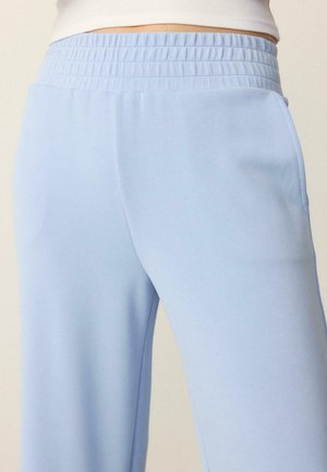 Približen posnetek osebe, ki nosi svetlo modre elastične hlače z višjim pasom in belo majico, prikazana je regija od pasu do sredine stegnenice.