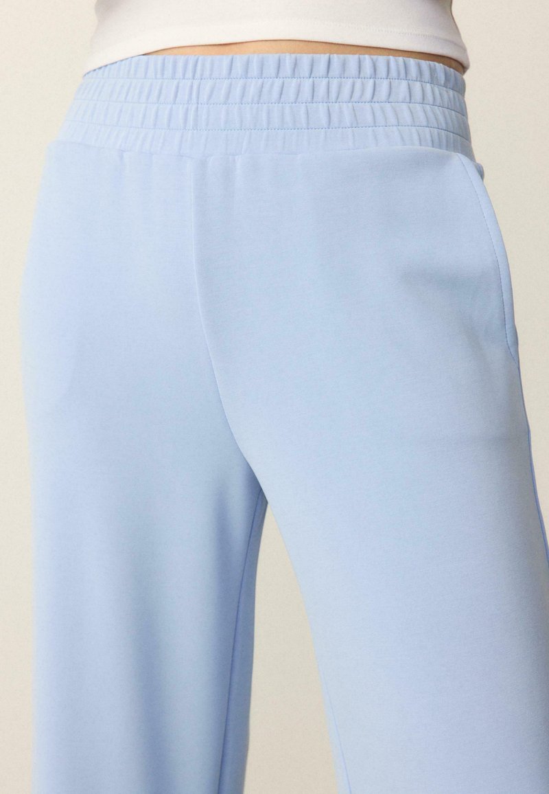 Gros plan sur une personne portant un pantalon à taille élastique bleu clair et un haut blanc, montrant la zone de la taille jusqu'au milieu des cuisses.