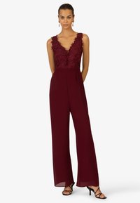 Burgunderfarbener Jumpsuit mit tiefem V-Ausschnitt, floralem Spitzenoberteil, weit geschnittenen Hosenbeinen und transparenten Details am Saum. Schwarze, römische Absatzschuhe.