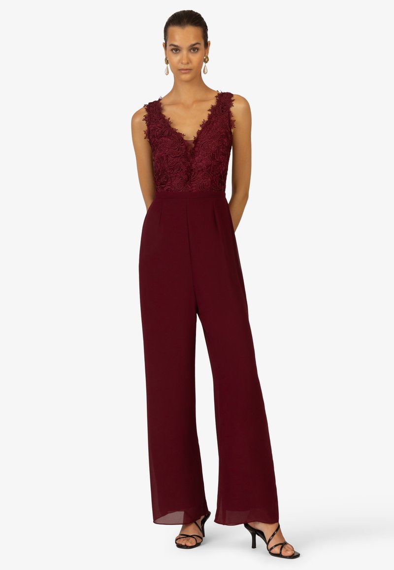 Burgunderfarbener Jumpsuit mit tiefem V-Ausschnitt, floralem Spitzenoberteil, weit geschnittenen Hosenbeinen und transparenten Details am Saum. Schwarze, römische Absatzschuhe.