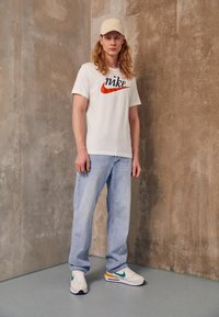 T-shirt bianca con logo "Nike" rosso e blu, jeans azzurri chiari e sneakers multicolori con accenti verdi e arancioni. Cappellino beige.