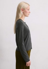 Gilet gris avec un col rond et une fermeture à boutons, associé à une jupe en velours côtelé verte. Le tissu semble doux avec une texture lisse.