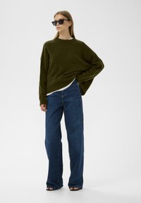 Olīvu zaļš pārpūsts džemperis ar strukturētu audumu, uzvilkts virs baltas blūzes, kombinēts ar platām tumši zilām biksēm un melnām saulesbrillēm.