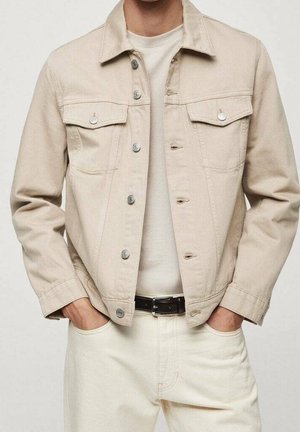 Mann trägt eine beige Jeansjacke über einem weißen Hemd mit weißen Hosen und schwarzem Gürtel, Hände in den Taschen.