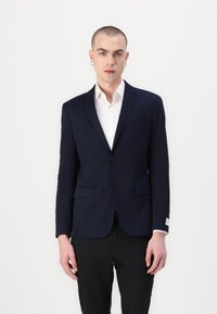 Marineblazer met een op maat gemaakt ontwerp, twee voorzakken, sluiting met één knoop en een subtiele, gestructureerde stof, gedragen over een witte blouse.