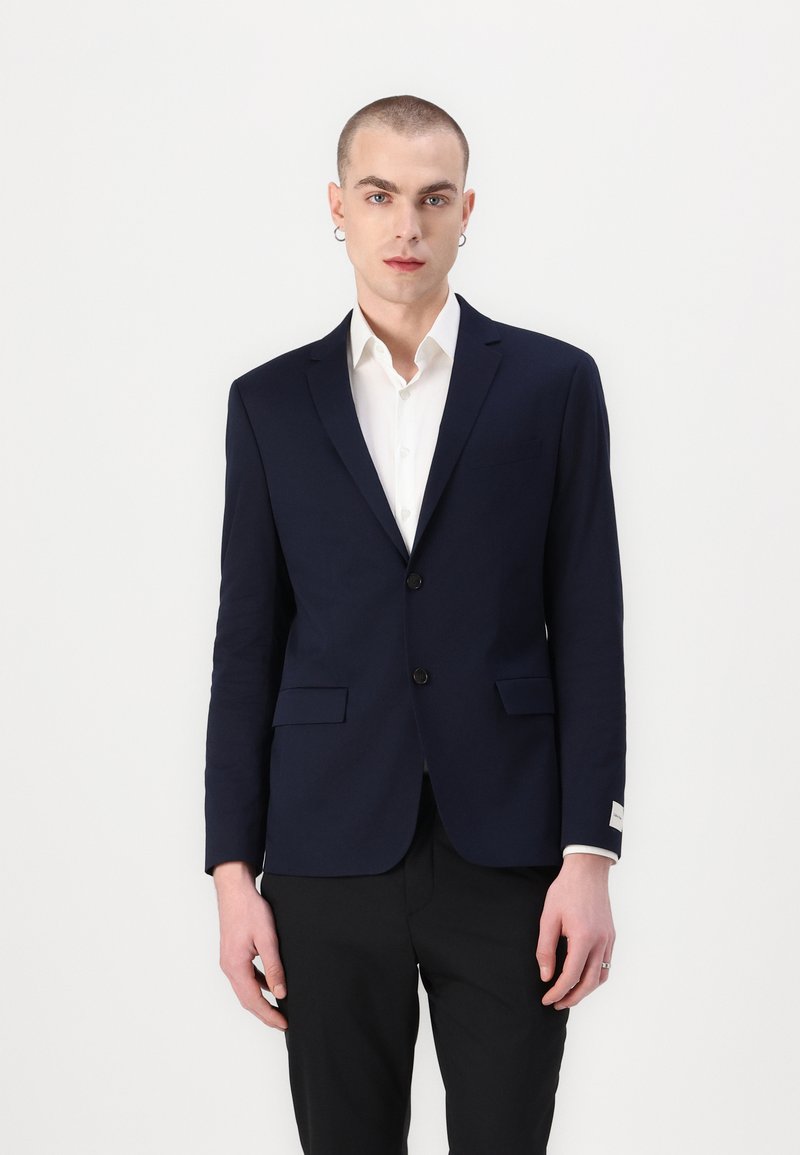 Marineblazer met een op maat gemaakt ontwerp, twee voorzakken, sluiting met één knoop en een subtiele, gestructureerde stof, gedragen over een witte blouse.