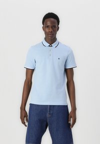 Lyseblå polo shirt med sort og hvid stribet krave, tre-knaps placket og lille logo på venstre bryst, parret med denimshorts.