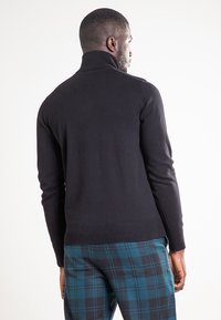 Czarny dzianinowy sweter z golfem z prążkowanymi mankietami i ściągaczem, zestawiony z niebiesko-czarnymi spodniami w kratę. Na swetrze nie ma widocznych wzorów.