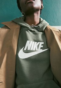 Grön huvtröja med en vit Nike-logotyp, kombinerad med en beige kappa. Har justerbar dragsko och texturerad tyg. Enkel, modern design.