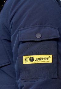 Manche d'une veste marine avec un patch aux accents jaunes portant l'inscription "JEANSCODE," indiquant la marque et la composition du tissu. Texture lisse, fermeture à bouton.