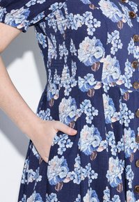 Námorně modré květované šaty s modrými a růžovými květy, s knoflíkovým uzávěrem, kapsou a texturovanými smockingovými detaily na živůtku.