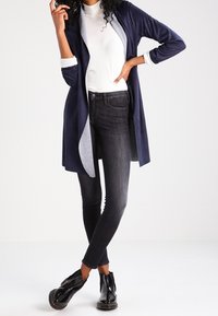 Marinblå lång cardigan över en vit polotröja, tillsammans med mörka skinny jeans och svarta ankelboots. Slät tyg med kontrasterande insida.