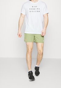 Camiseta blanca con el texto negro "NIKE RUNNING DIVISION", pantalones cortos verdes con un pequeño logo naranja y zapatillas deportivas negras.