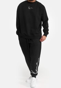 Felpa nera con logo bianco sul petto, abbinata a pantaloni della tuta neri con accenti bianchi lungo il lato. Sneakers bianche.