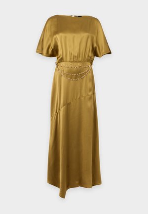 Femme aux cheveux bouclés portant une robe asymétrique dorée brillante avec une ceinture chaîne et des sandales à talons hauts noires avec bride à la cheville, debout sur fond blanc.