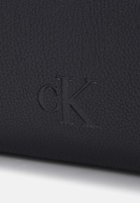 Bolso de mano de cuero negro con una superficie texturizada y un logo "cK" en relieve. El diseño es minimalista, sin hardware ni acentos adicionales.