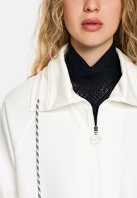 Weiße Zip-Jacke mit hohem Kragen, darunter ein strukturiertes schwarzes Spitzenoberteil und ein schwarz-weißes Kordelzug-Detail.