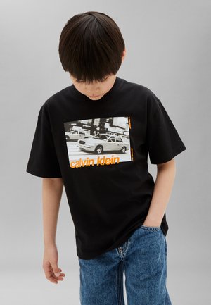 Dreng med mørkt hår iført sort Calvin Klein t-shirt med taxiprint og orange logo, kombineret med blå jeans, stående mod grå baggrund.