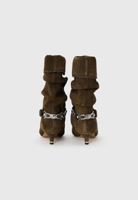 Stivaletti alla caviglia in suede marrone con un design arricciato, un dettaglio decorativo in catena color argento e un tacco sottile. Texture liscia.