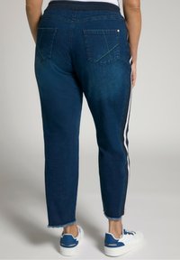 Joggers de mezclilla azul con cintura ajustada, bolsillos traseros con parche y rayas blancas laterales. Los dobladillos deshilachados y la textura suave completan el diseño.