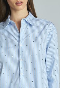 Camicia a righe blu chiaro e bianca con strass sparsi, dotata di colletto e maniche lunghe.