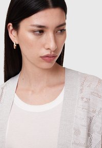 Cardigan lavorato a maglia grigio chiaro con apertura frontale e motivo simile a pizzo, indossato sopra una maglietta bianca a coste. Piccole orecchini ad anello dorati. Texture liscia.