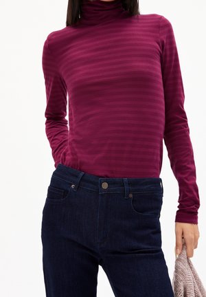 Kobieta ubrana w ciemnoróżowy, w paski golf i ciemnoniebieskie dżinsy, trzymająca prawą ręką beżowy, dziergany przedmiot.