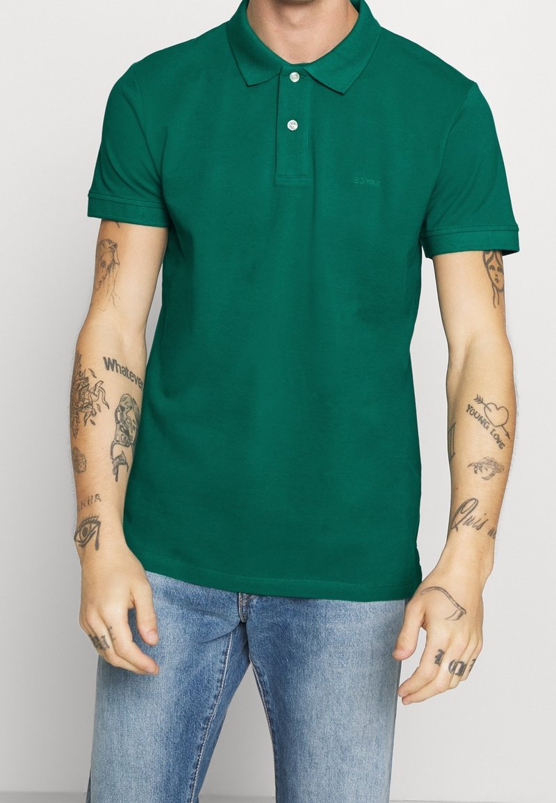 Polo en coton vert à manches courtes, patte à trois boutons et logo discret sur la poitrine ; accompagné d'un jean en denim bleu clair.