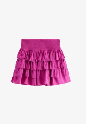 Smilende pige med krøllet hår iført hvid kort T-shirt med print af palme og papegøje samt lagdelt lyserødt nederdel med lyserøde sneakers.