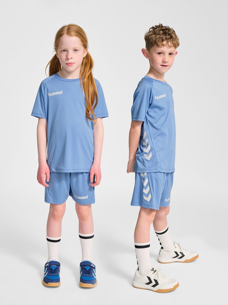 Deux enfants en tenue de sport bleue et chaussettes blanches se tiennent devant un fond uni, l'un fait face et l'autre est de profil.