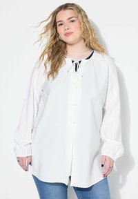Hvid langærmet bluse med rund halsudskæring, knaplukning foran og snøre detaljer ved halsen; har sammensunkne ærmer og en buet kant.