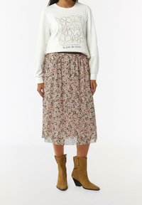 Kvinna iklädd en vit långärmad topp med blomsterdesign och text, blommig midikjol och beige mocka stövlar halvvägs upp på vaden med klackar.