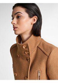 Abrigo marrón texturizado con un cuello alto, que cuenta con cuatro botones grandes y un bolsillo con cremallera lateral. Ajuste entallado con costuras pronunciadas.