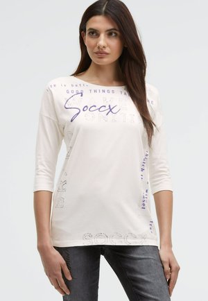 White cotton shirt with three-quarter sleeves, featuring purple printed text designs and a relaxed fit. Text includes phrases and brand name.