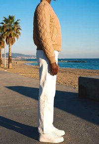Personne portant une veste matelassée beige, un pantalon blanc et des baskets blanches, debout sur une promenade de plage près des palmiers et de l'océan.