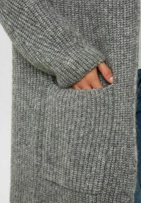 Grå stickad cardigan med ribbad textur, breda ärmar och en frampocket synlig på högra sidan. Mjuk, medelmjuk tyg.