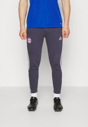Homme portant un pantalon de survêtement Adidas gris foncé avec des bandes blanches, le logo du Bayern Munich, des chaussettes blanches, des crampons noirs et un t-shirt bleu, visible de la poitrine aux pieds.