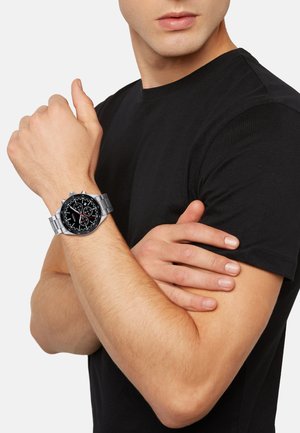 Silver stainless steel watch with a black dial, three sub-dials, and red accents, worn on a wrist with light skin tone and short nails.