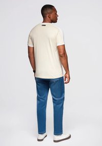 Beige tričko s krátkým rukávem, bílé akcenty, kulatý výstřih a malá černá nášivka. Nosené s modrými džíny a bílými teniskami.